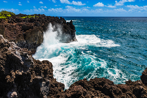 Large waves crashing against cliffs along the coastline of Maui on the road to Hana on a sunny day.