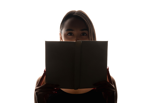 Reading mystery. Student fear. Exam anxiety. Dark backlit silhouette of scared woman eyes hiding face behind open book on white copy space background.