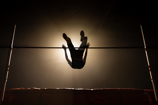 Sportswoman silhouette training high jump and flying through pole illuminated in dark gym wide shot