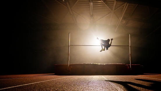 Sportsman silhouette training high jump and flying through pole illuminated in dark gym wide shot