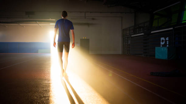 Sportsman walking at dark indoor stadium with sunlight shining through door back view wide shot
