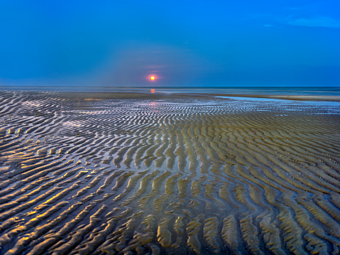 At low tide a red moon rises from the sea.