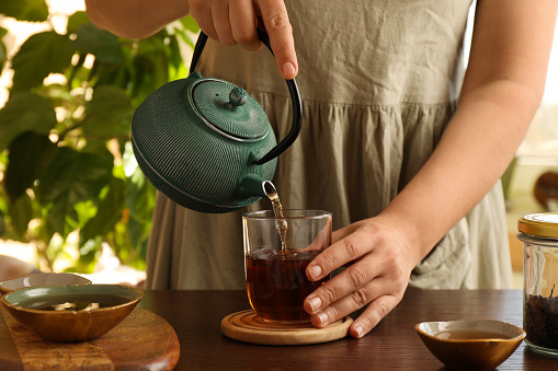 Woman pouring freshly brewed tea from teapot into cup at wooden table indoors, closeup. Traditional ceremony