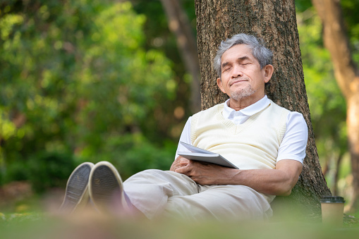 portrait healthy asian senior man closed eyes,siting against a tree on grasses while reading and relaxing in the park,concept of old people lifestyle,health benefits in  nature,wellbeing