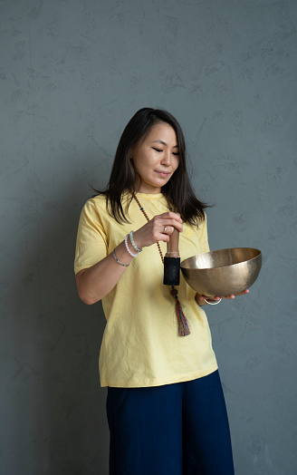 A young Asian woman holds a Tibetan singing bowl in her hands on a gray background with space for text.