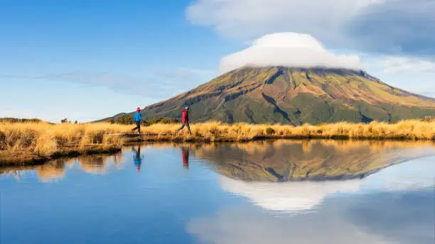 Father and son hiking Pouakai circuit with the reflection of  Mt Taranaki in the tarn in North Island, New Zealand.