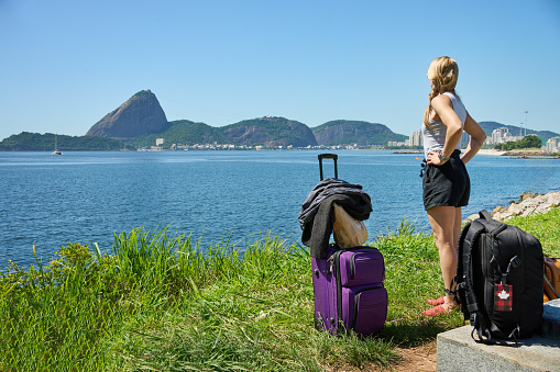 Rear view of a woman traveler with luggage standing at sea coast and looking at the seascape during summer