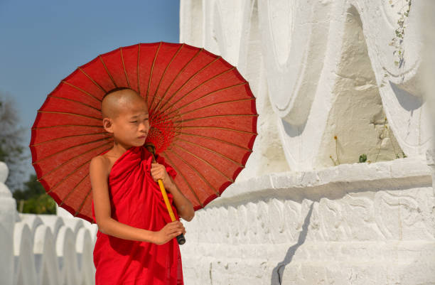 A Buddhist novice monk with a red umbrella walking at the white temple in sunny day.