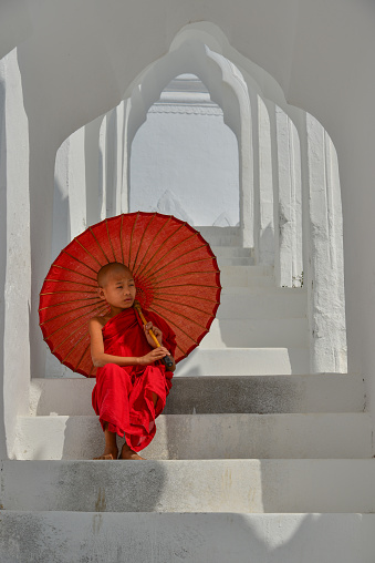 A Buddhist novice monk with a red umbrella sitting at the white temple.