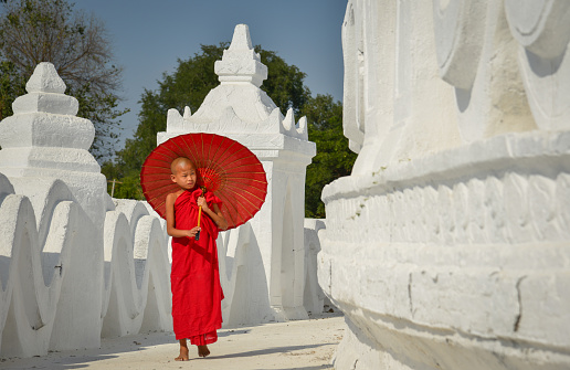 A Buddhist novice monk with a red umbrella walking at the white temple in sunny day.