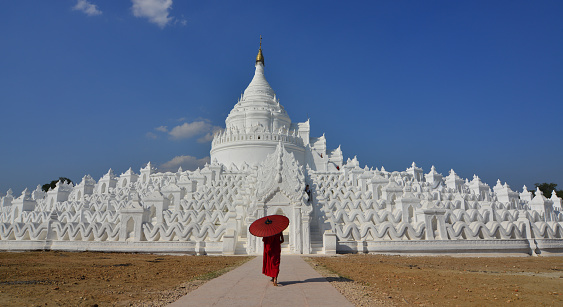 A Buddhist novice monk with a red umbrella coming to white stupa in sunny day.