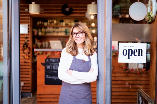 Portrait of confident female owner standing at entrance of cafe. Brunette haired beautiful woman looking at camera and smiling. Open sign in the door.