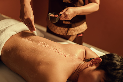 Masseuse hands pouring salt scrub on back customer at health spa. Relaxation man customer get service skincare scrubbing massage with masseuse in spa salon. Selective focus.