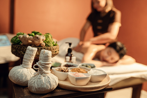 Selective focus, Herbal compress ball and healthy spa set on a table with customer relaxation massage on background. Spa and bodycare concept.