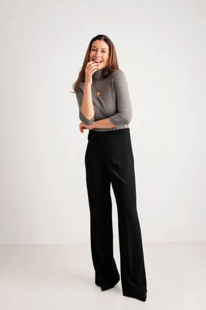 Full length of an attractive woman toothy smiling and looking at camera. Brunette haired female wearing turtleneck sweater and trousers while standing against isolated white background. Copy space.