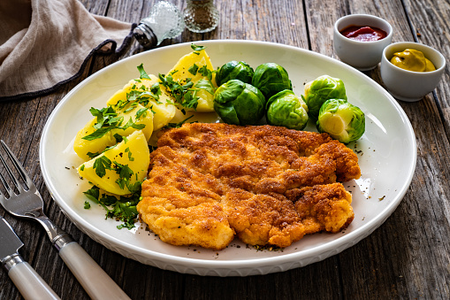 Breaded fried pork chop with boiled potatoes and boiled brussels on wooden table