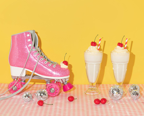 Summer creative layout with pink roller skate with whipped cream and bright red cherry, two milkshakes, disco balls, and cherries on pastel pink plaid and yellow background. 80s or 90s retro aesthetic idea. Minimal summer fashion girl idea.