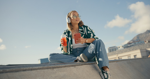 Fashion, music and woman with phone in city listening to audio, radio and track in skate park. Headphones, summer and female person relax outdoors on smartphone for social media, online and internet