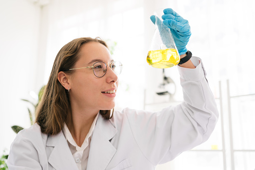 Caucasian woman scientist researcher wearing eye glasses shaking substance in the conical flask for analysis of liquids in the lab. Scientist working with a dropper and the conical flask.