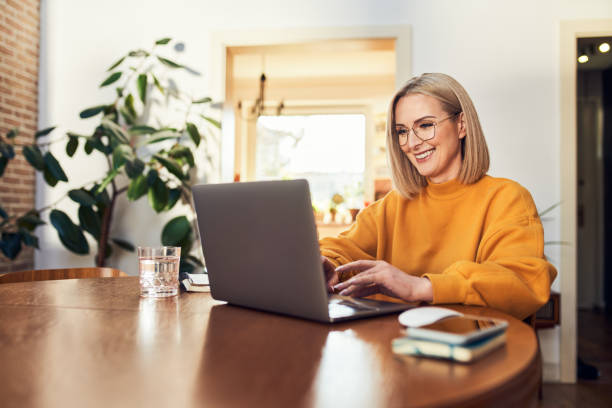 donna matura felice che utilizza il computer portatile mentre lavora in remoto da casa nel soggiorno - furniture internet adult blond hair foto e immagini stock