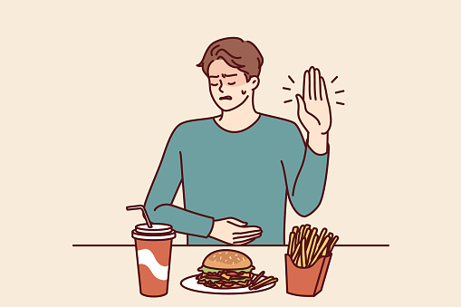 Man suffering from gastritis refuses fast food that causes pain in abdomen on advice of nutritionist. Guy near table with junk lunch shows stop gesture due to gastritis caused by eating hamburgers