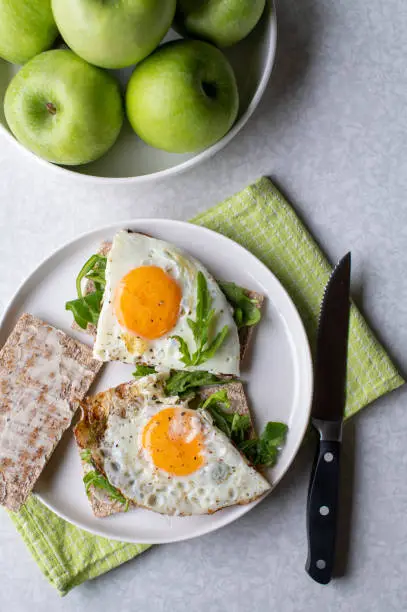 Healthy breakfast plate with fried egg, arugula sandwich. Served on buttered crispbread on light table background with green apples.
