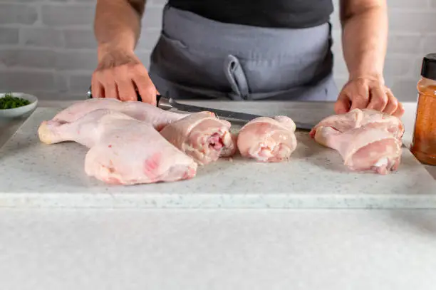 Woman with apron ist cutting or carving fresh and raw chicken legs into shanks and drumsticks on a cutting board. Closeup, front view