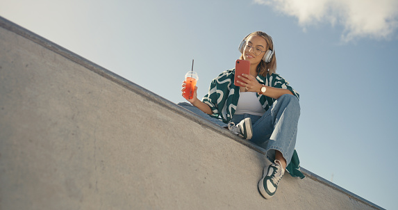 Phone, headphones and gen z girl on a rooftop with music, smoothie and texting from below. Freedom, smartphone and female student relax with streaming, podcast or radio, subscription or app outdoor