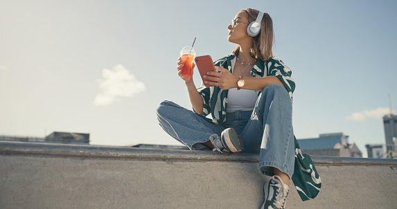 Woman, smartphone in city and drink, listening to music and looking at cityscape view, streaming and summer. Young female person relax in urban park, audio playlist and radio with ice tea and gen z