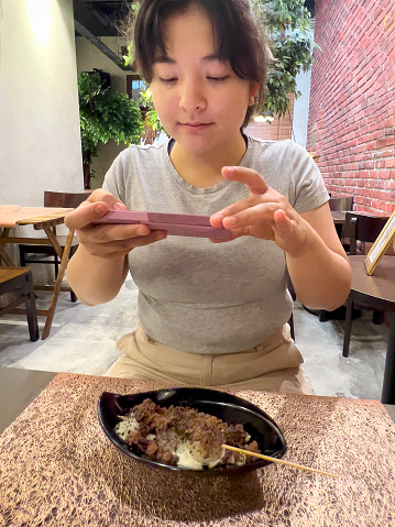 Eurasian young woman taking smartphone photo of chocolate, black sesame frozen glutinous ball desserts in a cafe.  George Town, Penang, Malaysia.