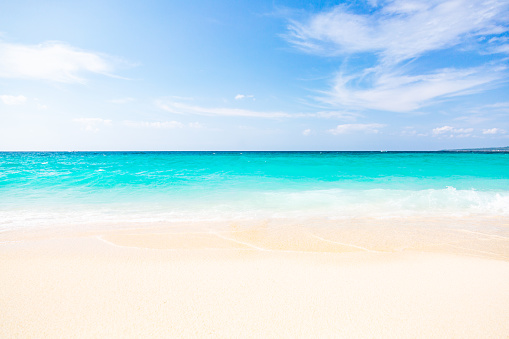 A scene from Puka Beach on Boracay Island, Philippines. The image portrays a sunny day at the beach, with a calm sea gently rolling onto the exceptionally clean, white sand shore. The water displays shades of turquoise and dark blue, gradually transitioning towards the horizon. The sky above appears blue with scattered clouds. The beach is empty, devoid of any people, creating a sense of tranquility and serenity.