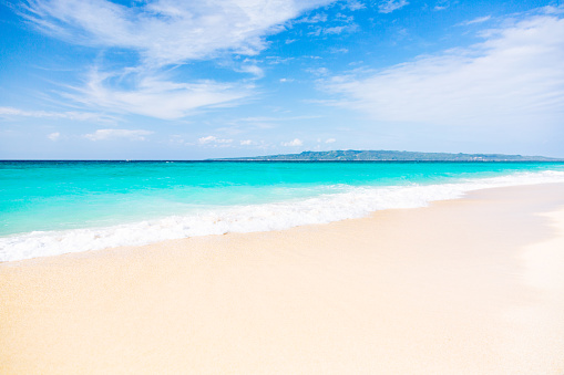 A scene from Puka Beach on Boracay Island, Philippines. A sunny day at the beach, with a calm sea gently rolling onto the exceptionally clean, white sand shore. The water exhibits shades of turquoise and dark blue, transitioning towards the horizon. In the distance, a small stripe of a hilly island can be seen. The sky above is blue, adorned with scattered clouds. The beach itself appears empty, devoid of any people, inviting a sense of tranquility and serenity.