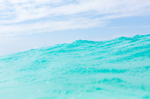 A closeup view of the sea surface, showcasing its remarkable characteristics. The water in the image exhibits a vibrant turquoise color and exceptional clarity. Ripples can be observed traversing the water, adding a sense of motion and texture to the scene. In the background, a blue sky is visible, adorned with scattered clouds that provide a visual contrast against the expanse of the sea.