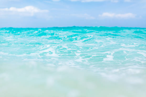 A surface of the sea after a wave crash, resulting in a foamy appearance. The water exhibits a vibrant turquoise color, while the background showcases a blue sky with clouds scattered throughout. The image captures the natural elements of the sea and sky, portraying the foam on the sea surface and the clouds in the sky.