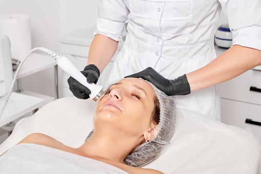 Cosmetologist makes ultrasound skin tightening for rejuvenation woman face using phonophoresis, anti aging cosmetic procedure with in beauty spa salon. Beautician makes ultrasonic skincare lifting