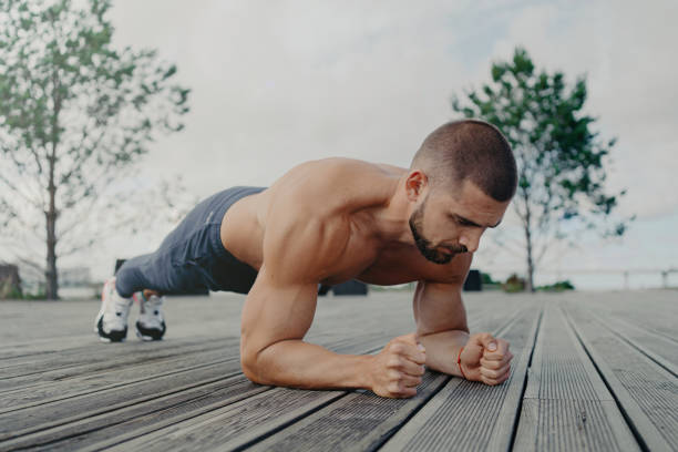 el atleta fuerte mantiene la postura de la plancha, priorizando la aptitud física y la salud. el hombre concentrado hace ejercicio al aire libre, enfatizando la fuerza abdominal. el culturista se centra en el entrenamiento central. - emphasizing fotografías e imágenes de stock