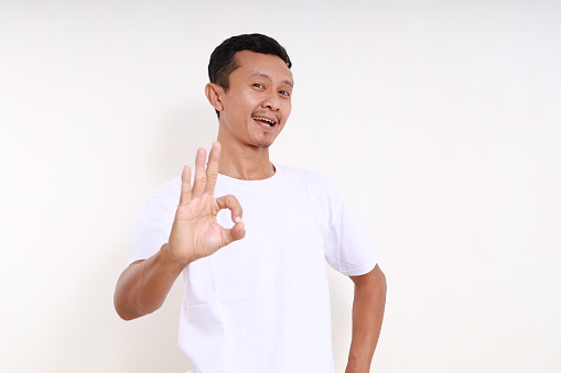 Happy asian funny man standing while showing okay hand gesture. Isolated on white background