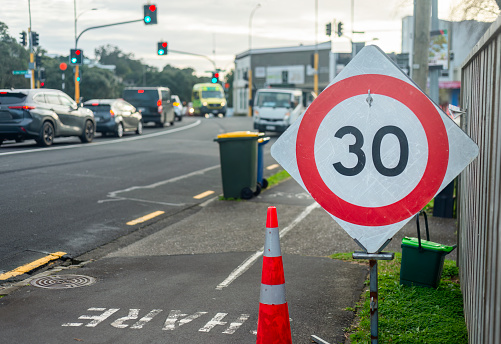 30km speed limit sign and rubbish bins by the roadside. Cars at intersection. Auckland.