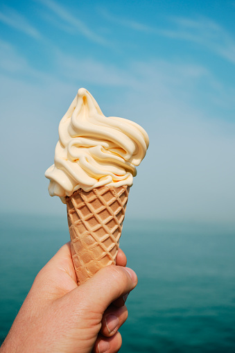 hand holding a delicious vanilla ice cream cone overlooking defocused sea at Newquay, Cornwall on a bright June day.