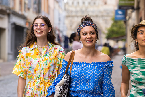 Waist-up shot of three young female adult friends walking through the town. They are all smiling, looking around, all wearing casual clothing. They are in the village of Moissac in the Southern region of France.