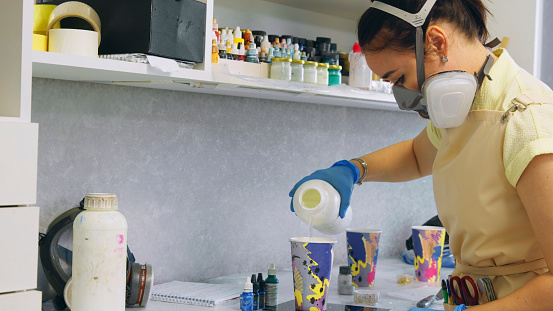 Wearing a protective face mask, the woman is pouring the material from the container into a disposable cup as part of her creative workshop tasks