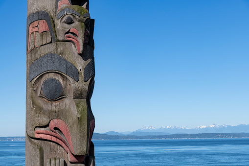 Seattle, WA, USA-July 2022; Close up view of part of a totem pole along the waterfront in Victor Steinbrueck Park with snow topped mountains of Olympic National Park on horizon