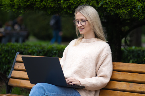 Young smiling blonde woman with glasses is sitting on a park bench with a laptop on her lap. Concept of freelancing, online communication, outdoor learning