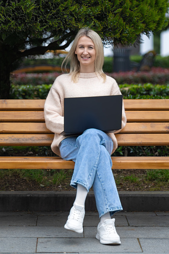 Una joven rubia feliz con una computadora portátil en el parque en un banco mira a la cámara. Chica estudiante, freelance, emprendedora en una computadora portátil en el parque. Foto vertical photo