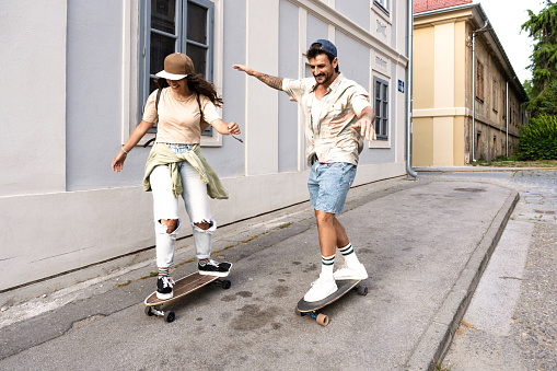 Tourists couple skateboarders riding skates. Happy young travelers longboarding together on sunset on pavement. Stylish man and woman in trendy outfit skating outdoors in touristic destination streets