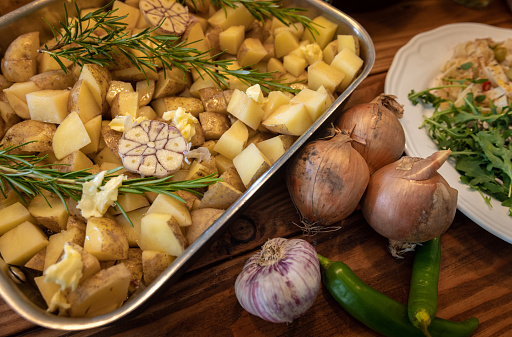 Close-up of a cooking tray filled with sliced potato, garlic, rosemary and butter on a wooden table ready to cook. On the table there is also a garlic clove, onions and green chilli peppers.