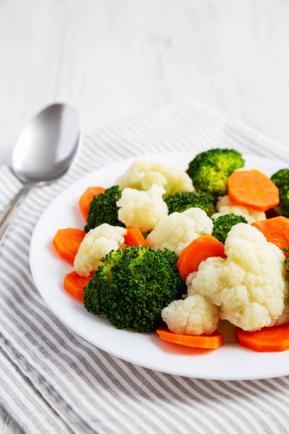 istock Mixed Organic Steamed Vegetables (Carrots, Broccoli and Cauliflower) on a Plate on a white wooden background, side view. 1502877996