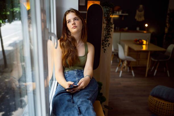 携帯電話を持ちながら窓越しに見る物思いにふける白人の10代の少女 - teenager adolescence portrait pensive ストックフォトと画像
