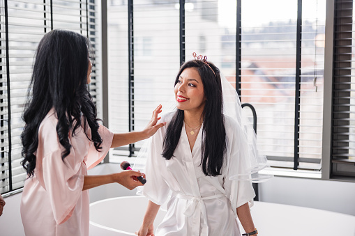An engaged Latin female getting her makeup done by her best friend before they go out to celebrate her bachelorette. They are located in luxurious bathroom wearing silk bathrobes. They are smiling and having fun catching up and having fun conversations.