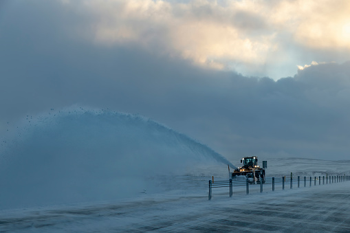 Driver’s perspective view over main road around Iceland covered in snow with snowplow on other side of fenced median throwing snow on side against a dark snow cloud with some bright light and blue sky